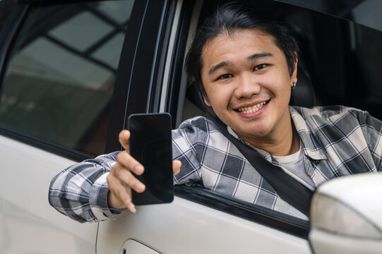 Asian man smiling from car window, wearing plaid shirt and seatbelt, cheerfully showing a smartphone screen toward the camera, symbolizing connectivity or mobile service - Powered by Adobe