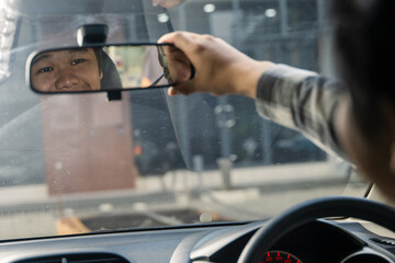 Asian man adjusting car rearview mirror while smiling, reflection showing his face, viewed from behind the steering wheel inside the vehicle on a bright day
