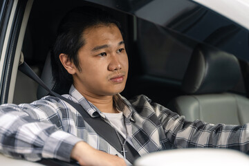 Asian man with serious expression sitting behind the wheel inside a car, wearing a plaid shirt and...