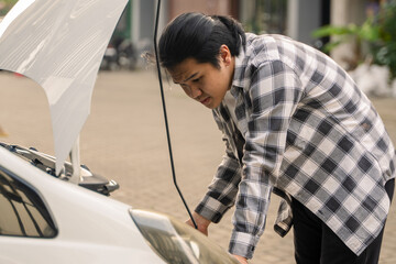 Asian man inspecting the engine under the open hood of a white car, looking concerned and leaning...