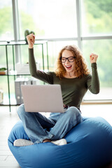 Young woman celebrating success while working on a laptop in a comfortable indoor space with a...