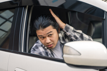 Asian man sitting in a car, adjusting his hair with one hand while looking down with a focused...