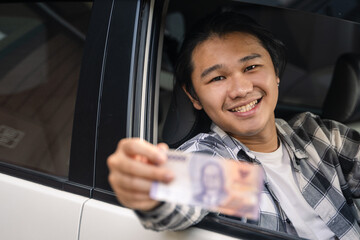 Asian man smiling while sitting in a car, holding out Indonesian banknote from the window,...