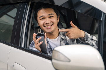 Asian man inside a car smiling while pointing at an unbuckled seatbelt, promoting safety awareness...