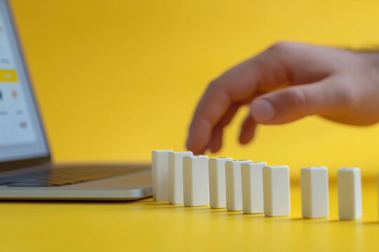 Business, Technology, and Risk - Hand Pushing Dominoes Next to Laptop hand dominoes risk laptop work