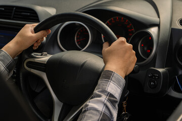 Close-up of an Asian person’s hands holding the steering wheel of a car from the driver’s seat,...