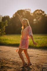 Smiling woman in a colorful hippie dress with headband dances joyfully in the sunlit countryside,...