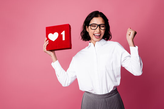 Charming businesswoman celebrating success holding a love sign box on a pink background wearing glasses and white shirt - Powered by Adobe