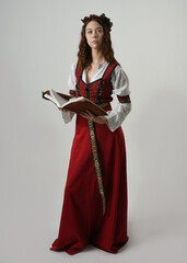 Fototapeta premium portrait pretty young female brunette model wearing a red medieval gown, historical fantasy costume. holding a book, reading pose. Isolated figure on studio background