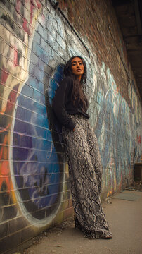 Young South Asian woman in her 20s posing confidently in urban alley, wearing a red crop top and python print pants, leaning on graffiti wall. Perfect for fashion editorials and youth culture blogs
