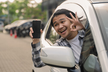Asian man smiling and holding a black smartphone out of a car window while parked in an outdoor area during the daytime