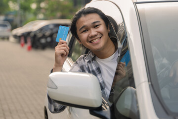 Asian man smiling while showing a blue credit card out of the car window in an outdoor parking area...