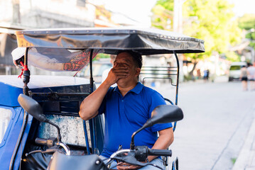 Middle-aged Southeast Asian tricycle driver in blue shirt covers his mouth as he yawns tiredly...