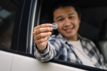 Asian man sitting in a car, smiling while holding a silver 500 coin outside the window with focus...