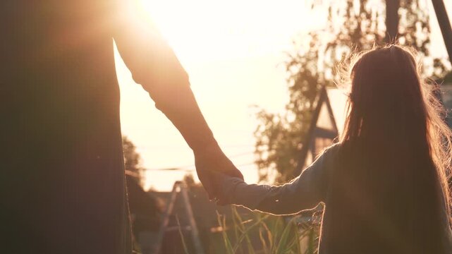 Warm sunlight illuminates mother and daughter holding hands while walking in park at sunset. Warm evening, joint outdoor walks. Bond between mother and child. Silhouette peoples in bright sunlight