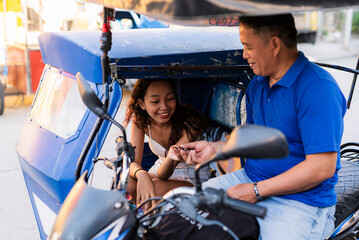 A cheerful Southeast Asian tricycle driver receives payment from a smiling young woman, sharing a friendly and positive street moment.