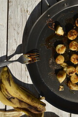 Fried banana slices in caramel on black frying pan on white wooden background. Preparation process
