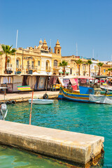 Fototapeta premium Malta Republic, Malta Island. 28. 06.2023. Marsaxlokk city, The harbour and traditional coloured boats on a hot sunny day. This town has a big market every weekend.