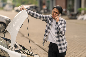 Asian man calling for help while standing beside a white car with the hood open, showing a...