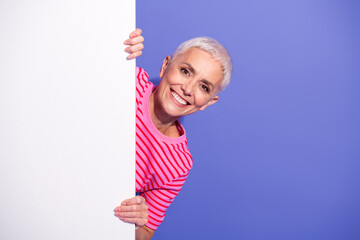 Charming mature woman in a striped shirt cheerfully peeking over a white board against a vibrant...