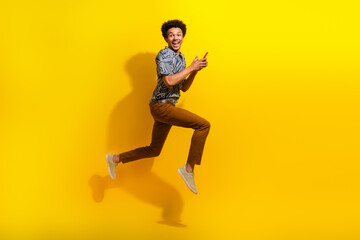 Excited young man in stylish outfit happily leaping with phone against vibrant yellow background,...
