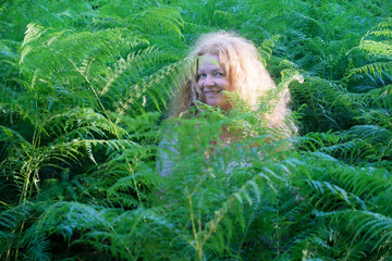 Joyful woman with long curly ginger red hair peeks out from dense green ferns, her bright smile and...