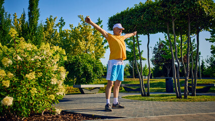 Older man stretching arms in park under morning sun in yellow shirt and shorts. Concept of morning routine, flexibility, healthy aging, outdoor wellness, mental clarity.