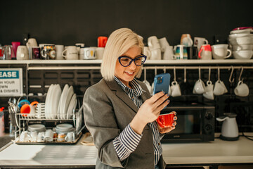 Businesswoman using smartphone and drinking coffee during break in office kitchen