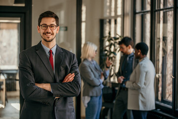 Confident businessman is posing in modern office while his colleagues are discussing in background
