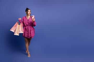 Young woman in stylish pink dress holding shopping bags and a smartphone against a vibrant purple...