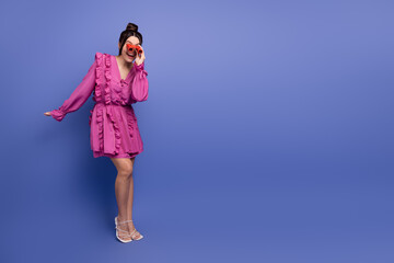 Charming young woman in a pink ruffled dress posing playfully on a purple background with stylish...