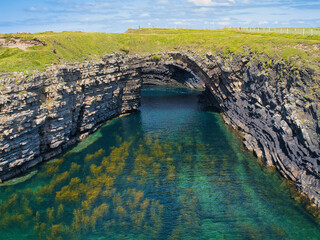 Stunning rock strata arch over vivid turquoise waters, enhanced by lush green surroundings at the Bridges of Ross, Ireland.