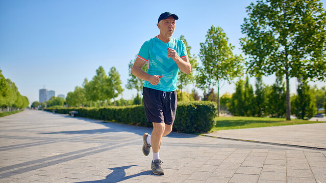 Older man running outdoors on sunny day in urban park. Concept of senior fitness, summer workouts, health promotion, active lifestyle, awareness for aging well.