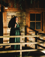A woman in an emerald satin dress stands on a rustic wooden porch bathed in dramatic light and deep...