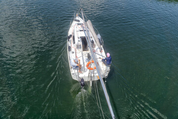A sailboat maneuvers powered by the engine without sails, with the mast lowered, aerial view. Two sailors on board. Leasure, travelling, vacations.