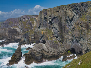 Dramatic cliffs rise against a clear blue sky at Mizen Head, Ireland, with waves crashing below. Captures rugged coastal beauty.