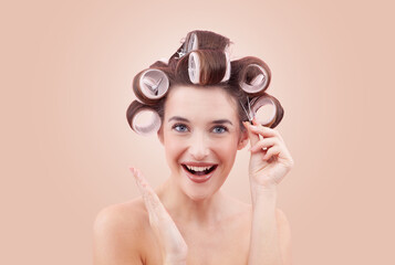 Portrait of a smiling young woman with a hairpin and hair curlers in her hair. Happy girl applying rollers while looking at the camera. Hairstyle, beauty, and self-care. Isolated on beige background