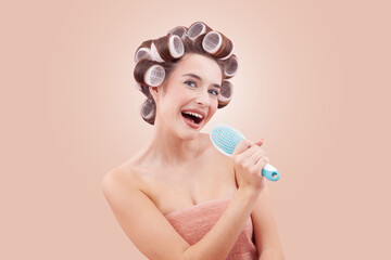 Portrait of smiling young woman with hair curlers, holding a hairbrush like a microphone and sing. Happy girl enjoying her beauty routine. Hairstyle, beauty and self-care. Isolated on beige background