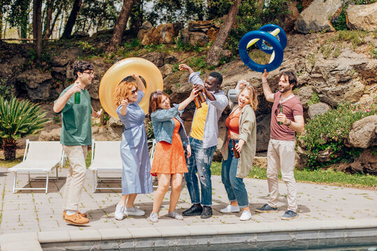 Friends toasting with beer bottles and dancing by the poolside at a summer pool party