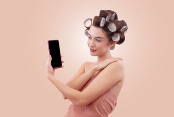 Woman with hair curlers and holding a smartphone. Concept of hairstyle, beauty routine, and self-care. Shopping online or contacting a hairdresser for an appointment. Isolated on beige background.