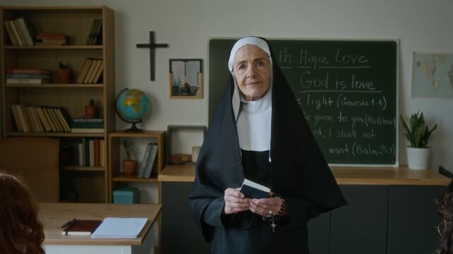 Medium slowmo portrait of mature Caucasian nun in veil posing for camera holding Bible and rosary in well-equipped classroom in modern religious school