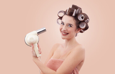 Portrait of a smiling young woman with hair curlers, holding a hair dryer. She enjoying her beauty routine after a bath or shower. Personal care and hairstyle concept. Isolated on beige background.