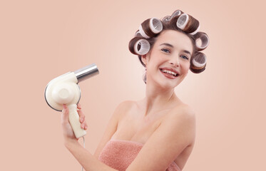 Portrait of a smiling young woman with hair curlers, holding a hair dryer. She enjoying her beauty routine after a bath or shower. Personal care and hairstyle concept. Isolated on beige background.