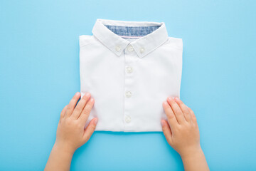Baby boy hands holding new folded white shirt on light blue table background. Pastel color....
