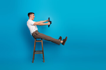 Young man pretending to drive seated on a stool against vibrant blue backdrop showcasing casual...