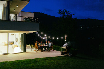 A group of young diverse people having dinner on the terrace of a modern house in the evening. Fun for friends and family. Celebration of holidays, weddings with barbecue