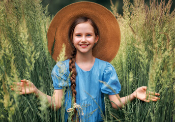 Portrait of a girl kid in a straw hat is standing in in a wheat field in summer.