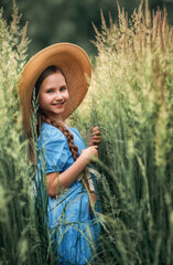 Portrait of a girl kid in a straw hat is standing in in a wheat field in summer.