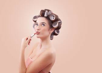 Make up, young woman with hair curlers applying lipstick on kissing lips while looking to the side with her blue eyes. Concept of hairstyle, makeup, beauty routine and cosmetics. Isolated on beige.