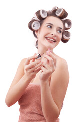 Young smiling woman with hair curlers applying nail polish while looking to the side with her blue eyes. Concept of hairstyle, makeup, beauty routine, and cosmetics. Isolated on white background